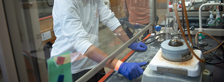 Person with gloved hands using lab equipment behind plexi shield